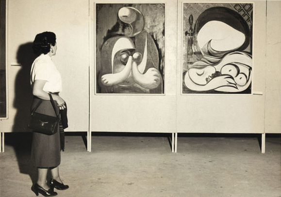 Judith Lauand observa as obras <i>Figura em Cadeira Vermelha</i> e <i>O Espelho</i> de Pablo Picasso durante a 2ª Bienal de São Paulo